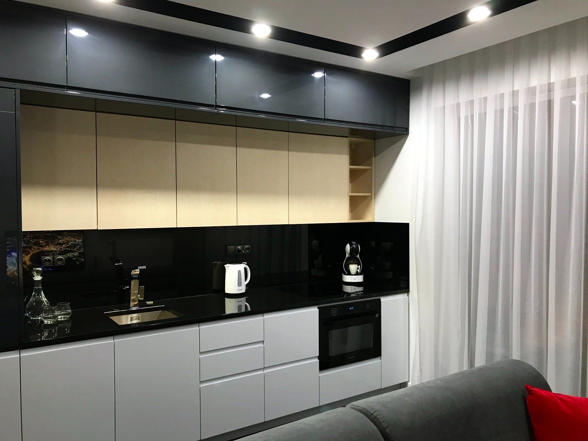 Nowoczesna kuchnia z szarymi szafkami dolnymi, czarnymi szafkami górnymi i czarnym blatem, uzupełniona o sprzęt AGD i dekoracyjne naczynia, widziana z perspektywy salonu.