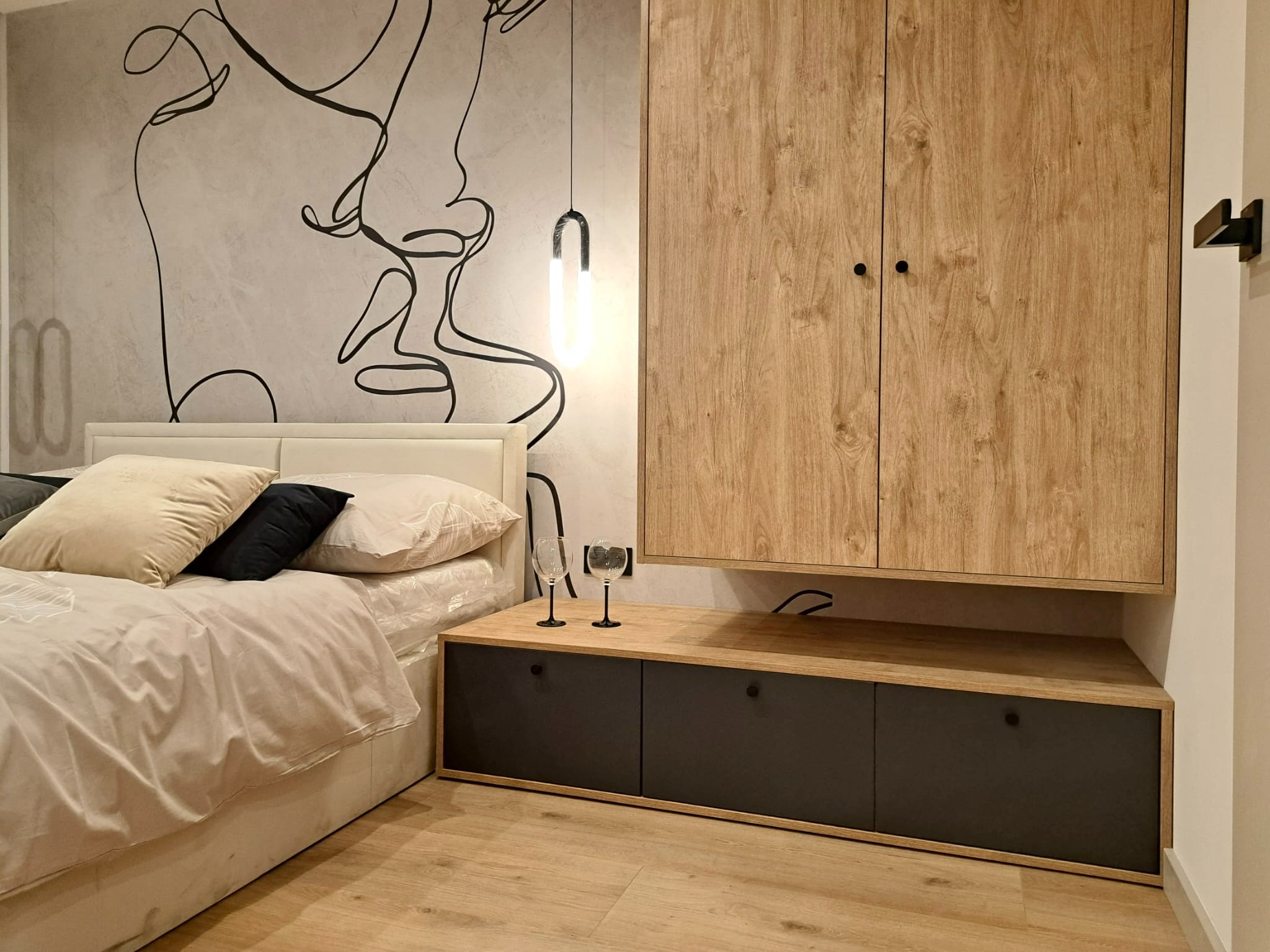 Nowoczesna sypialnia z drewnianą szafą wiszącą, szafką nocną z szufladami, łóżkiem z beżowym zagłówkiem i abstrakcyjnym rysunkiem na ścianie.