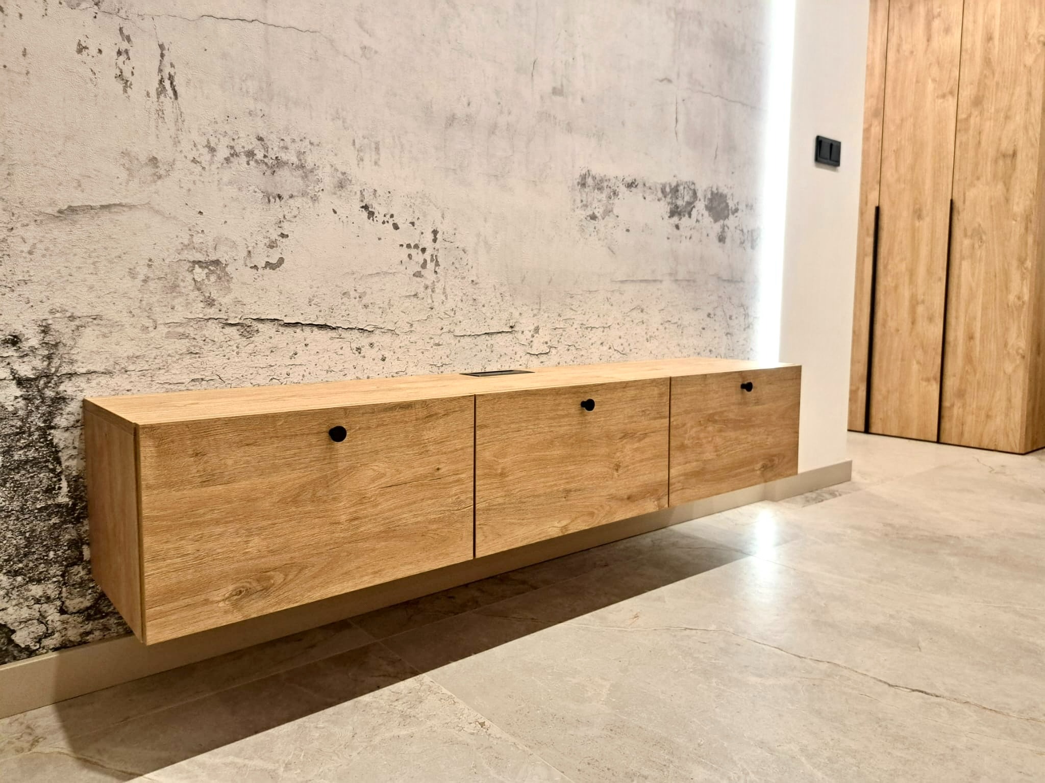 Wisząca szafka RTV z drewnianymi frontami na tle ściany o surowym wykończeniu, widoczne czarne uchwyty i fragment szafy wnękowej z identycznego materiału.