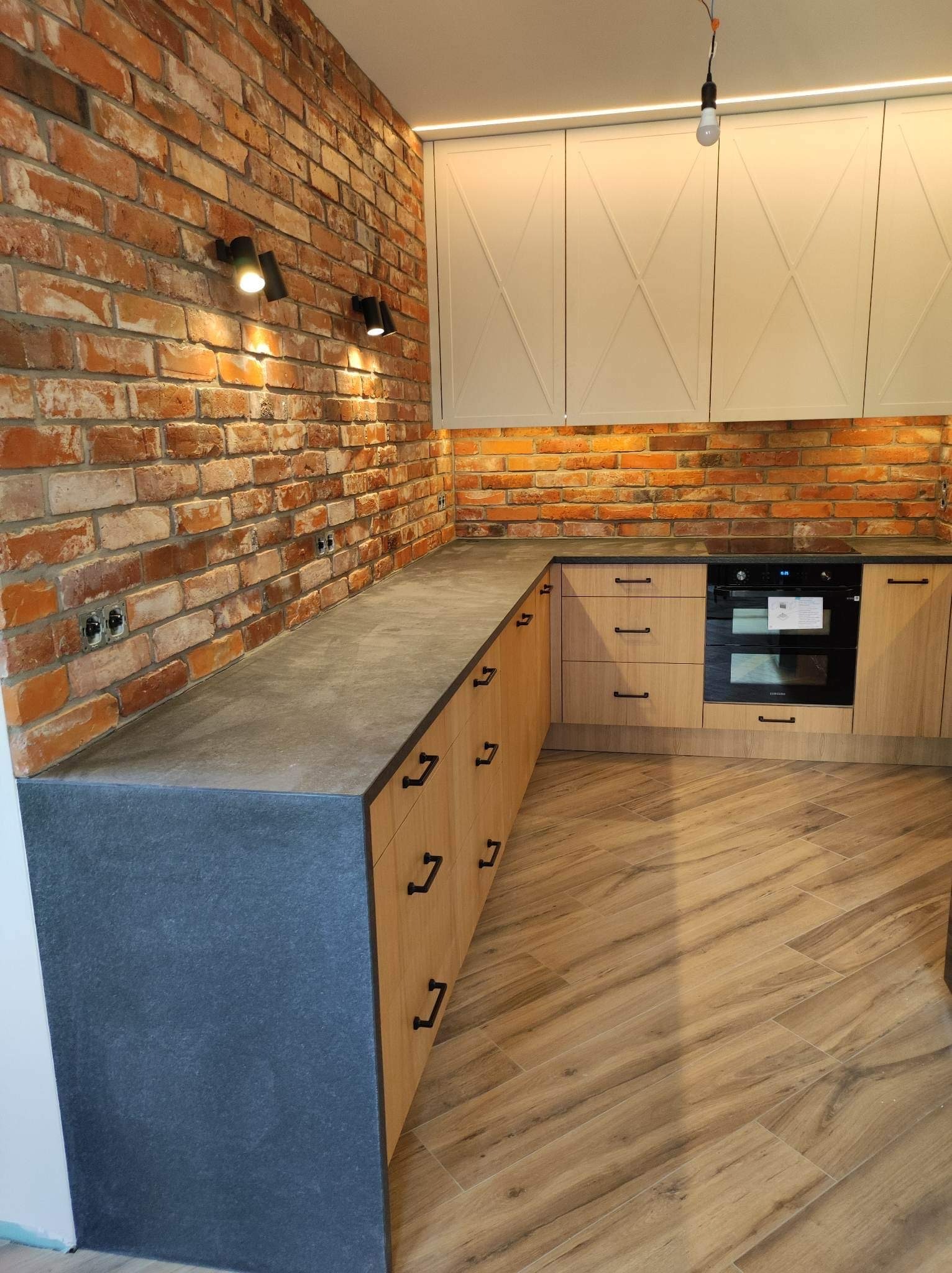 Nowoczesna kuchnia z ceglaną ścianą, szarym blatem i drewnianymi frontami szafek z czarnymi uchwytami. Widoczna zabudowa kuchenna z piekarnikiem i płytą grzewczą.