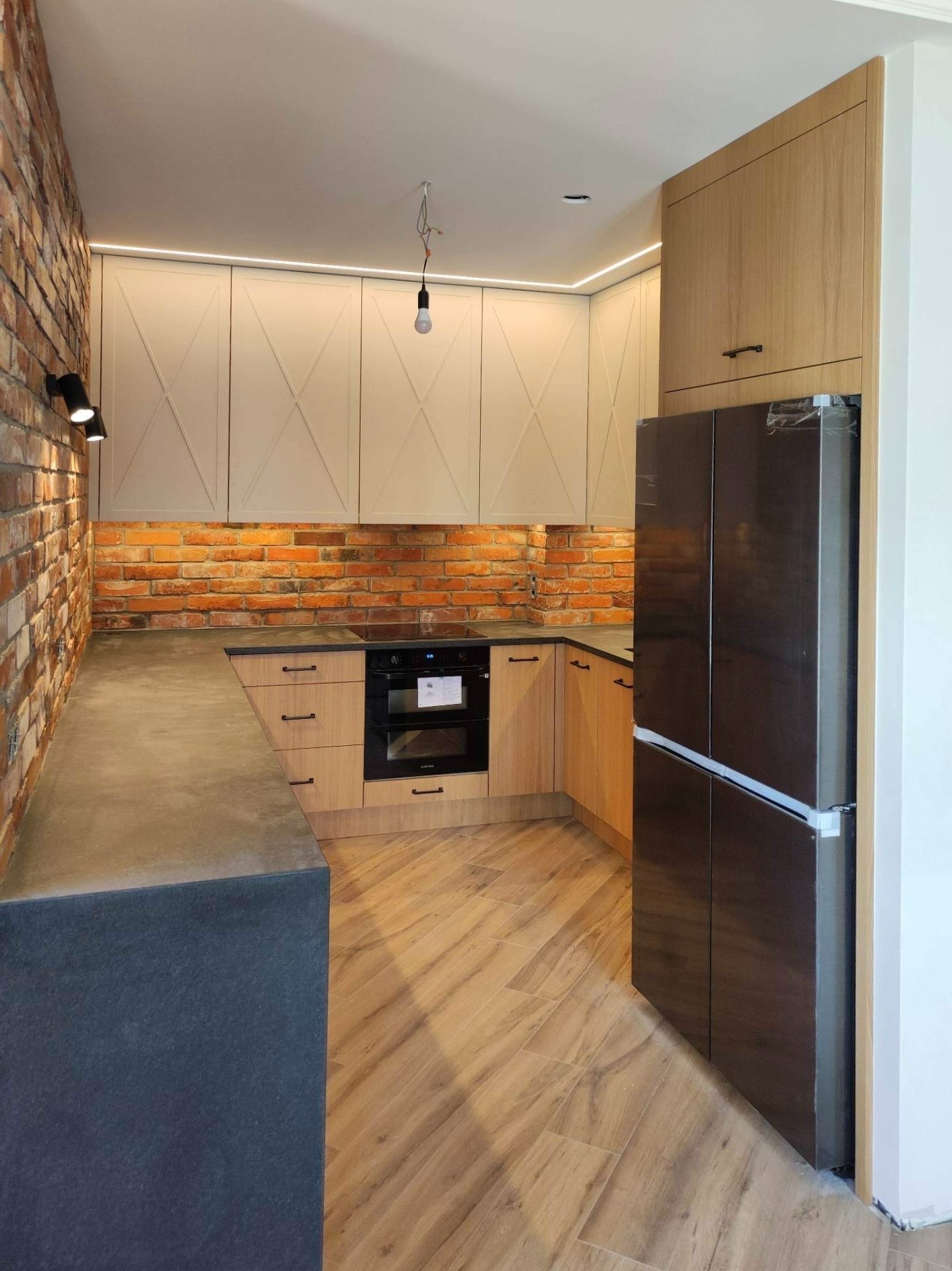 Nowoczesna kuchnia z ceglaną ścianą, drewnianymi frontami szafek z krzyżowym wzorem, blatem w kolorze grafitu i nową, jeszcze ofoliowaną lodówką.