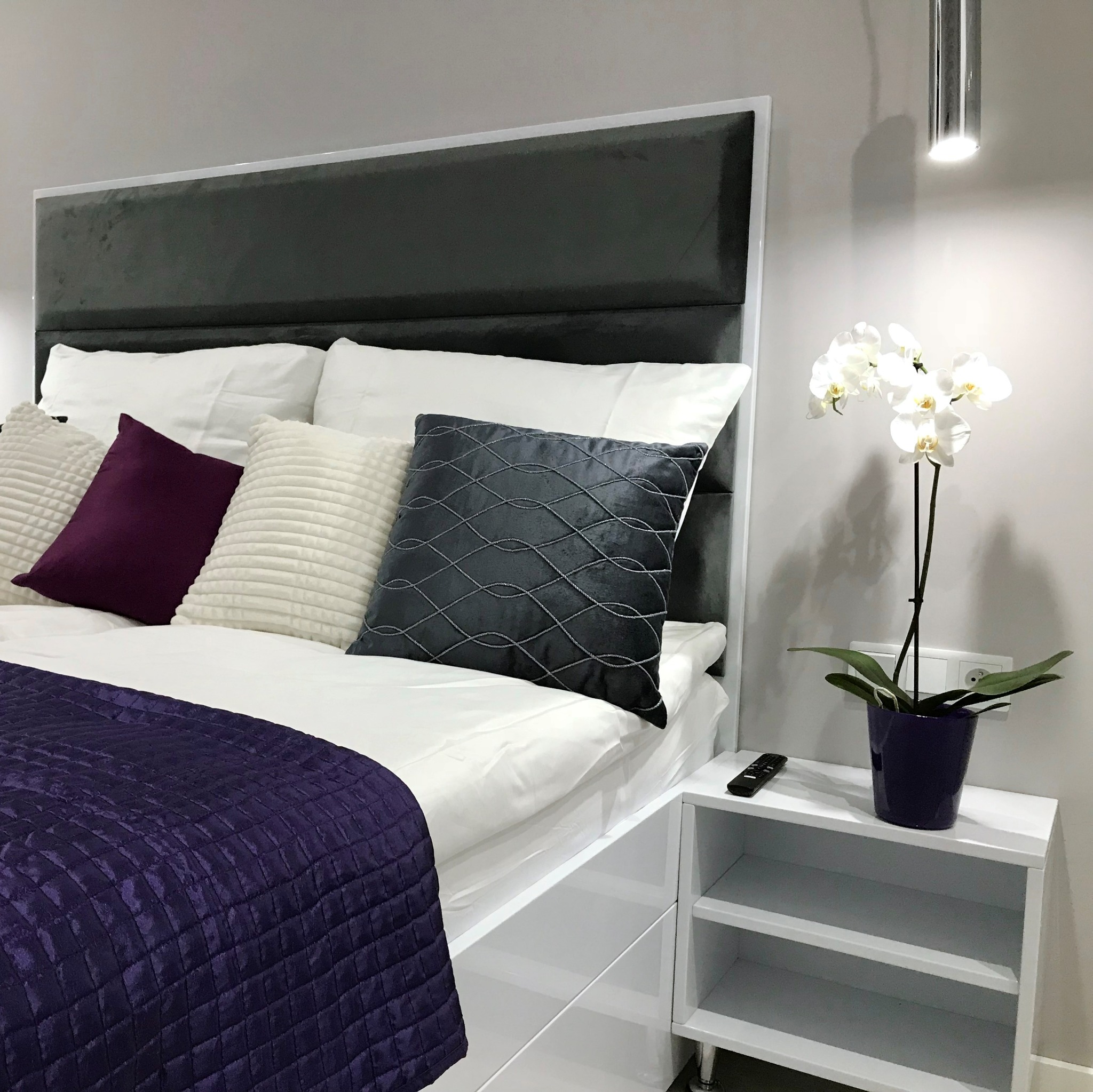 Nowoczesna sypialnia w Mysłowicach z tapicerowanym zagłówkiem w kolorze grafitowym, białą pościelą, dekoracyjnymi poduszkami w odcieniach fioletu i szarości oraz białą szafką nocną z orchideą...
