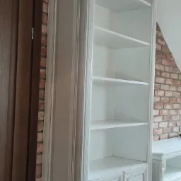 Biała, wysoka szafa wnękowa z półkami i dolnymi drzwiczkami, z dekoracyjnymi elementami, stojąca na drewnianej podłodze przy ścianie z czerwonej cegły.