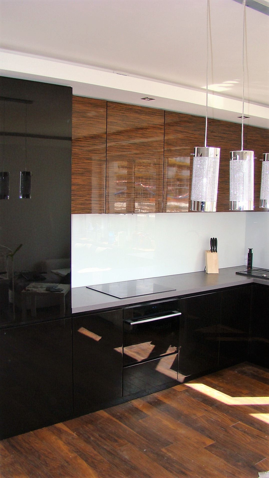 Nowoczesna kuchnia z czarnymi, lakierowanymi frontami szafek dolnych i górnymi szafkami z fornirem w odcieniach brązu, oświetlona trzema wiszącymi lampami z kryształkami, z drewnianą podłogą...