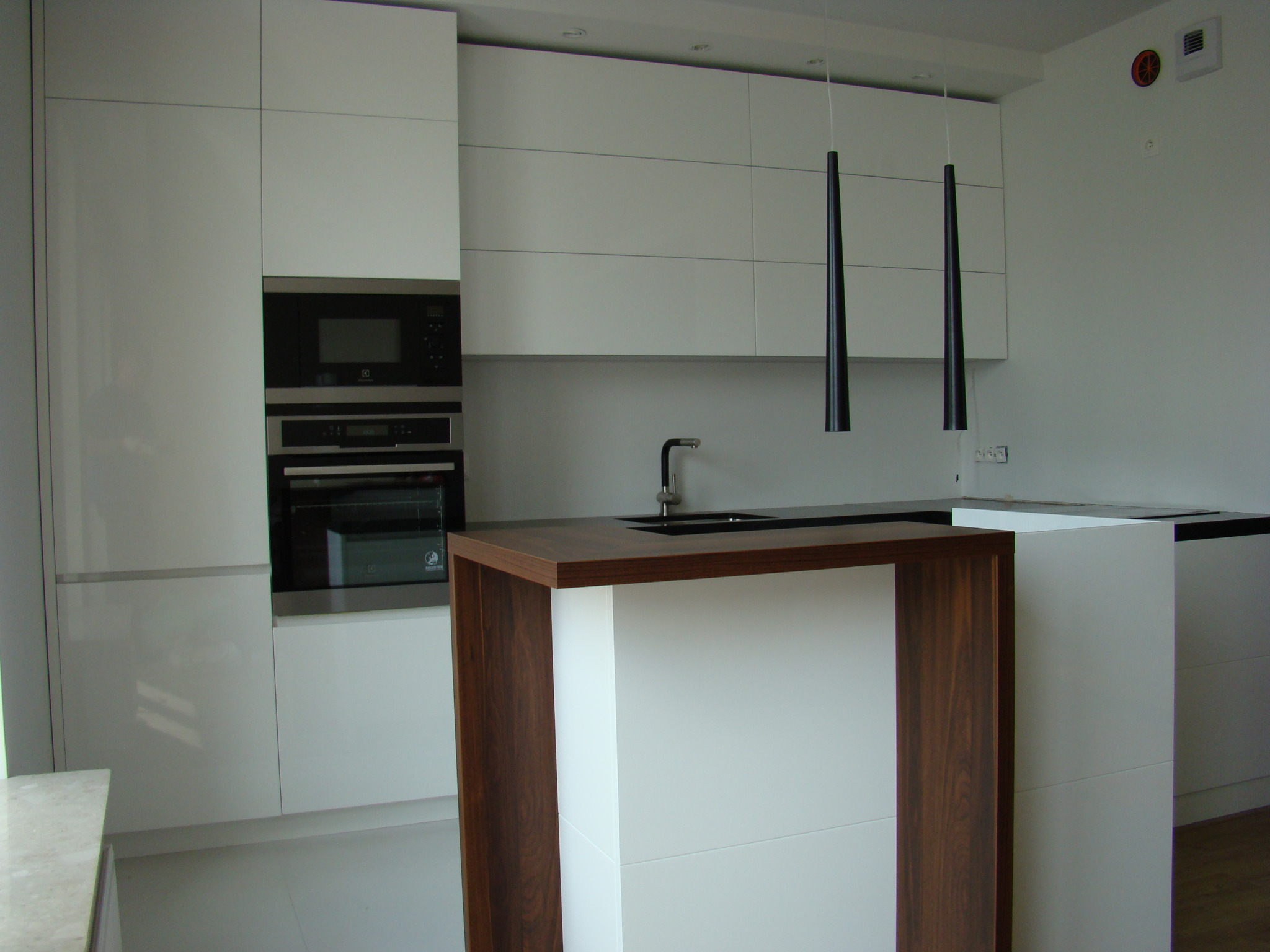 Minimalistyczna, biała kuchnia z wyspą o drewnianym obramowaniu i czarnym blatem, oświetlona dwoma wiszącymi, stożkowymi lampami.