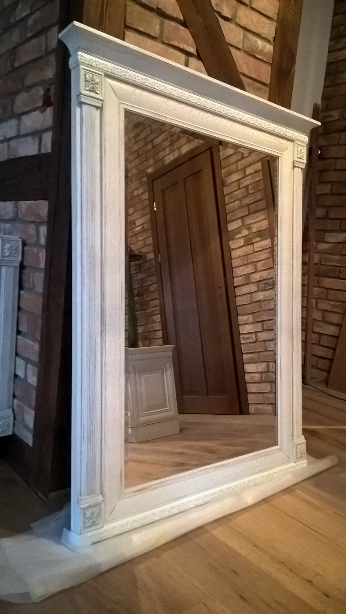 Ozdobna, kremowa rama lustra z dekoracyjnymi detalami, stojąca na drewnianej podłodze, z ceglaną ścianą i drewnianymi belkami w tle, odbijająca drzwi i fragment pomieszczenia.
