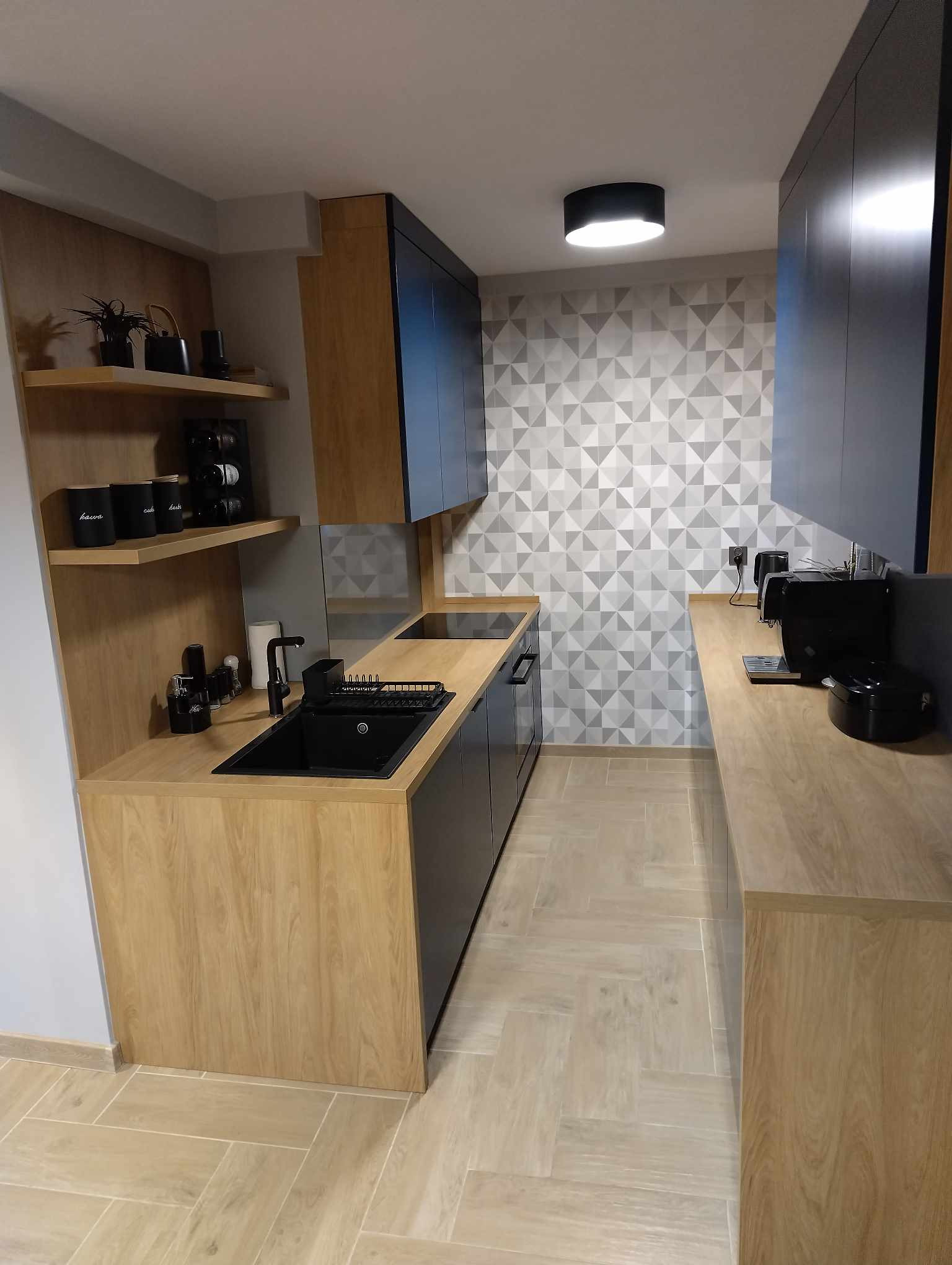 Wnętrze kuchni z drewnianymi blatami, czarnym zlewem i szafkami w kolorze granatowym, uzupełnione geometryczną tapetą i drewnianymi półkami z dekoracjami.