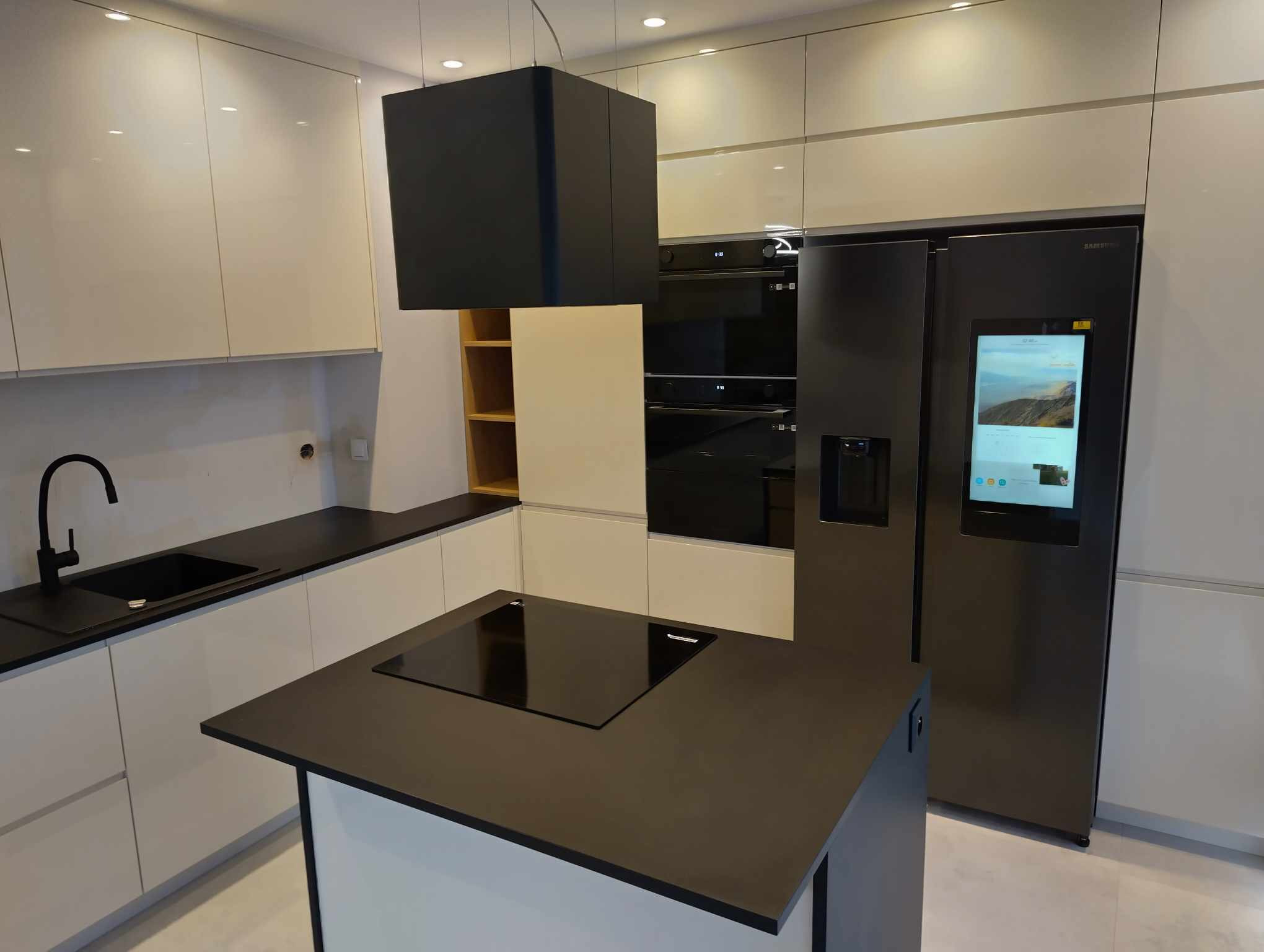 Nowoczesna kuchnia z wyspą, białe szafki z połyskiem, czarny blat, czarna armatura, zabudowany sprzęt AGD, lodówka Samsung z ekranem.