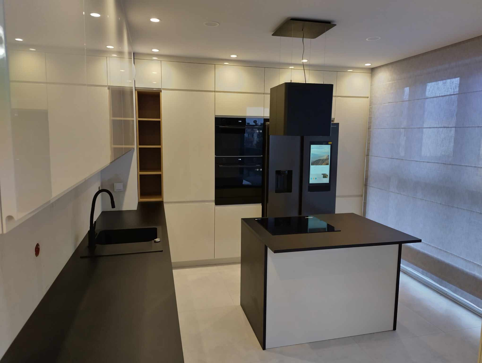Nowoczesna kuchnia z białymi, lakierowanymi frontami szafek, czarnym blatem i wyspą kuchenną z płytą indukcyjną, widoczny również czarny zlew i bateria oraz inteligentna lodówka z ekranem.