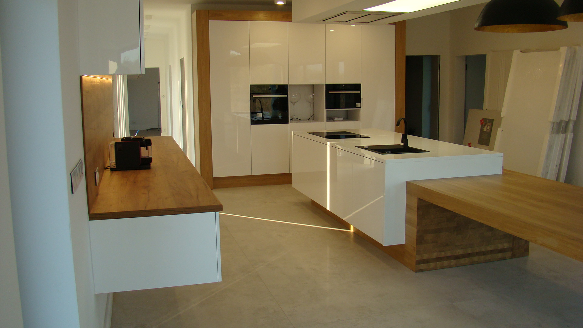 Nowoczesna kuchnia z białymi, lakierowanymi frontami, drewnianym blatem i wyspą kuchenną z czarnymi detalami, oświetlona światłem dziennym.