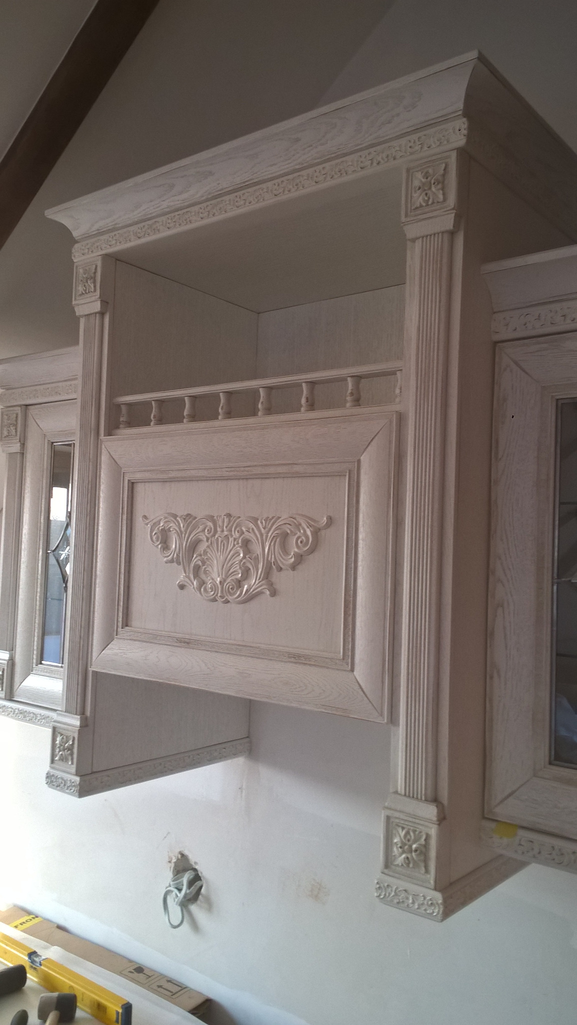 Klasyczna, kremowa szafka kuchenna z ozdobnymi listwami i ornamentem, widoczne narzędzia na blacie poniżej, sugerujące montaż.