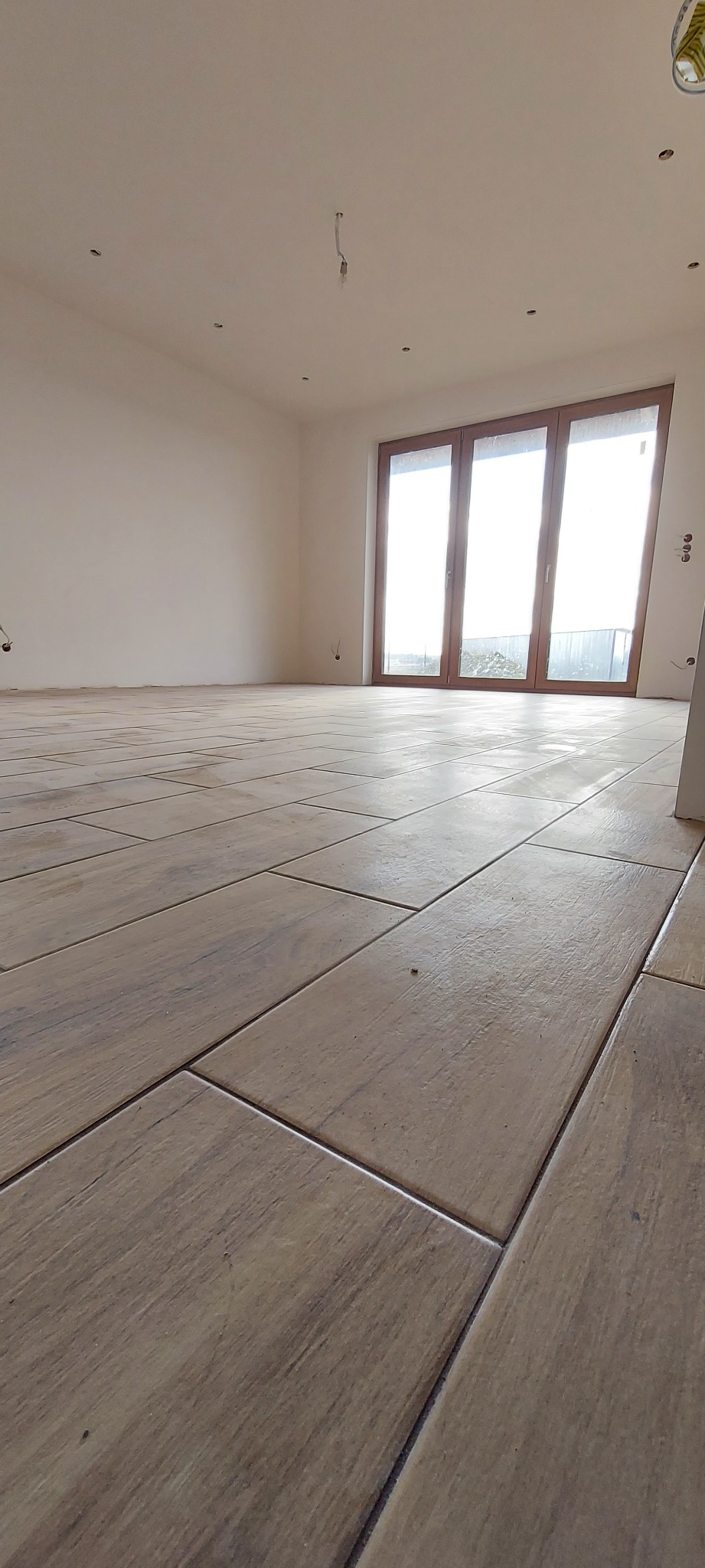 Salon w stanie deweloperskim z nową podłogą z płytek imitujących drewno, widoczne niedokończone instalacje elektryczne na ścianach i suficie, duże okno balkonowe z widokiem na zewnątrz.