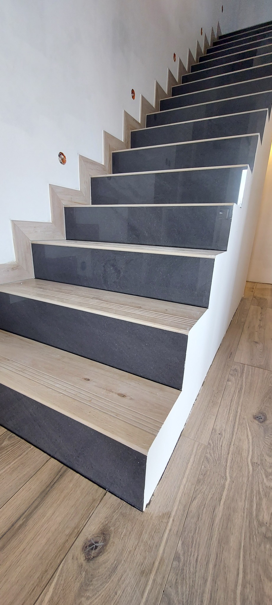 Schody z drewnianymi stopnicami i ciemnoszarymi podstopnicami, wykończone białą listwą przyścienną, widoczne z dołu, z drewnianą podłogą na pierwszym planie.