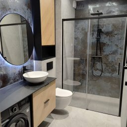 PROJBUD Agnieszka Czyżniewska - Nowoczesna łazienka z umywalką nablatową, okrągłym lustrem z podświetleniem LED, kabiną prysznicową typu walk-in z czarną armaturą i pralką w zabudowie pod blatem.