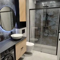 Nowoczesna łazienka z umywalką nablatową, okrągłym lustrem z podświetleniem LED, kabiną prysznicową typu walk-in z czarną armaturą i pralką w zabudowie pod blatem.