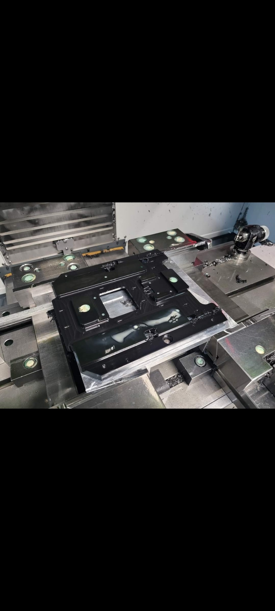 Czarny element z tworzywa sztucznego, prawdopodobnie obudowa, zamocowany na stole frezarki CNC, widoczne ślady chłodziwa i opiłki po obróbce.