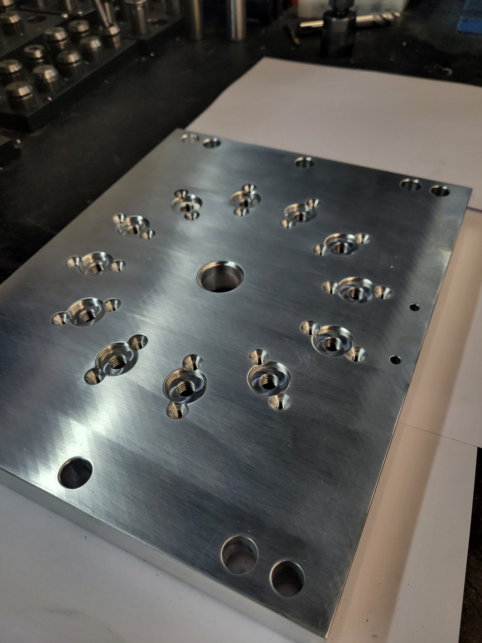 Szlifowana aluminiowa płyta z precyzyjnie wywierconymi i nagwintowanymi otworami o różnych średnicach, przygotowana do montażu podzespołów, leżąca na białym papierze na warsztatowym stole.