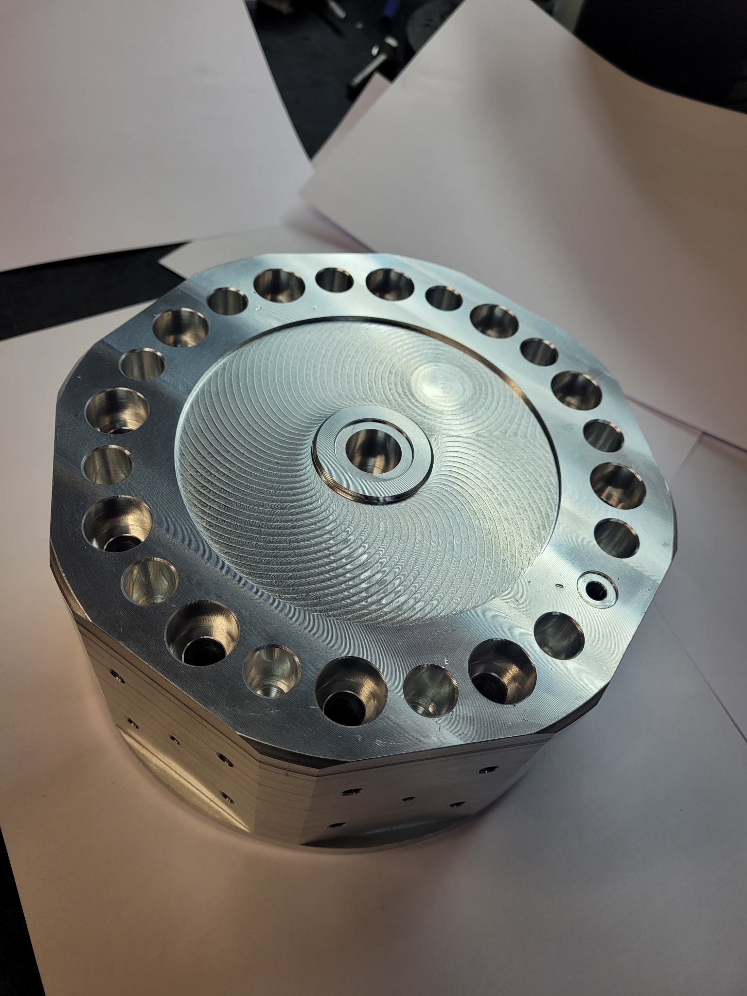 Precyzyjnie obrobiony element z metalu o okrągłym kształcie z koncentrycznymi rowkami i otworami na obwodzie, ułożony na białym papierze.