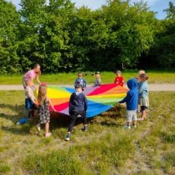 Grupa dzieci i dorosły trzymają wspólnie dużą, okrągłą chustę w kolorach tęczy na trawiastym terenie z drzewami w tle.