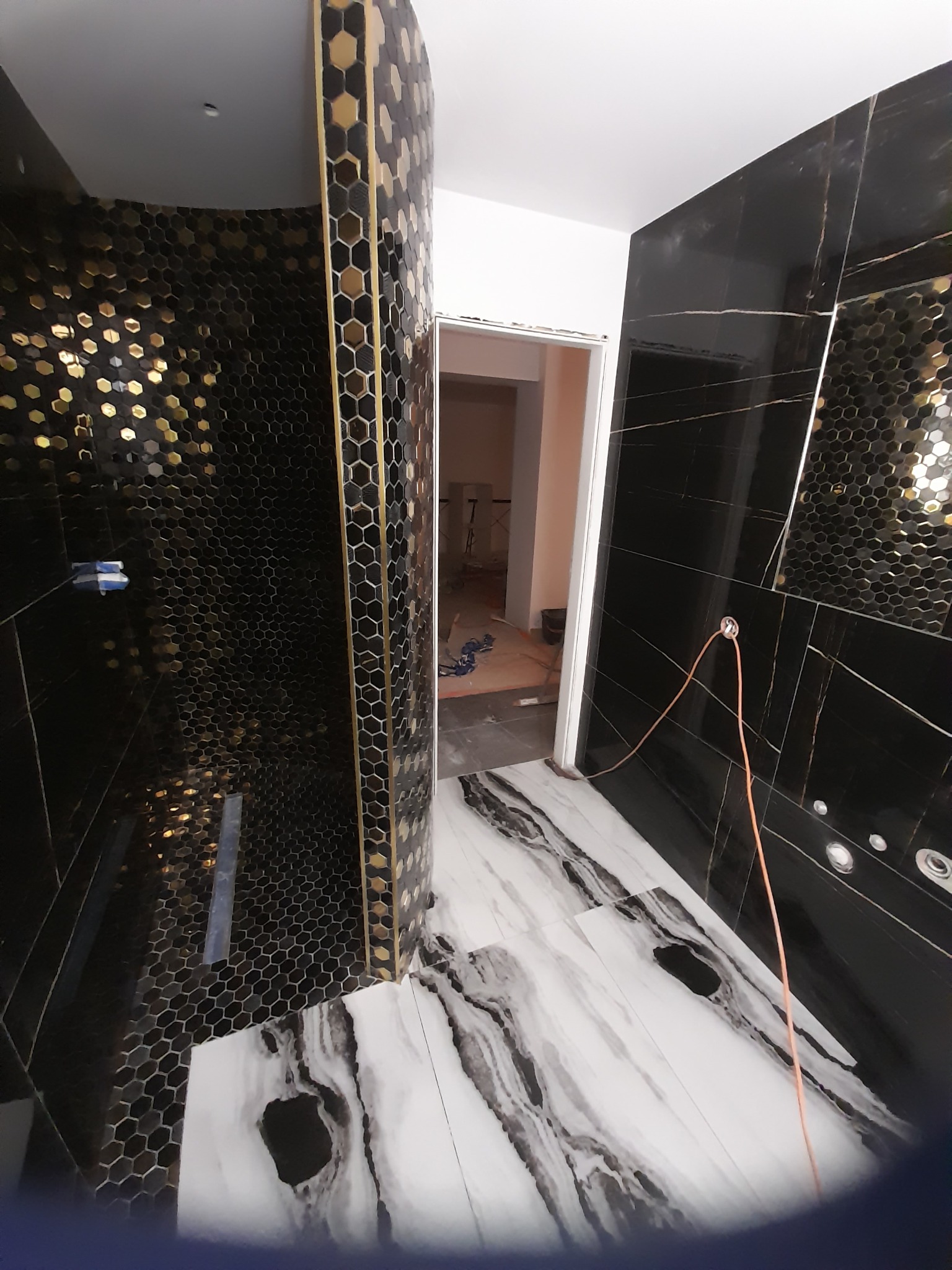 Częściowo wykończona łazienka z czarnymi płytkami imitującymi marmur na ścianach i heksagonalną mozaiką w strefie prysznica, biała podłoga z szarymi żyłami.