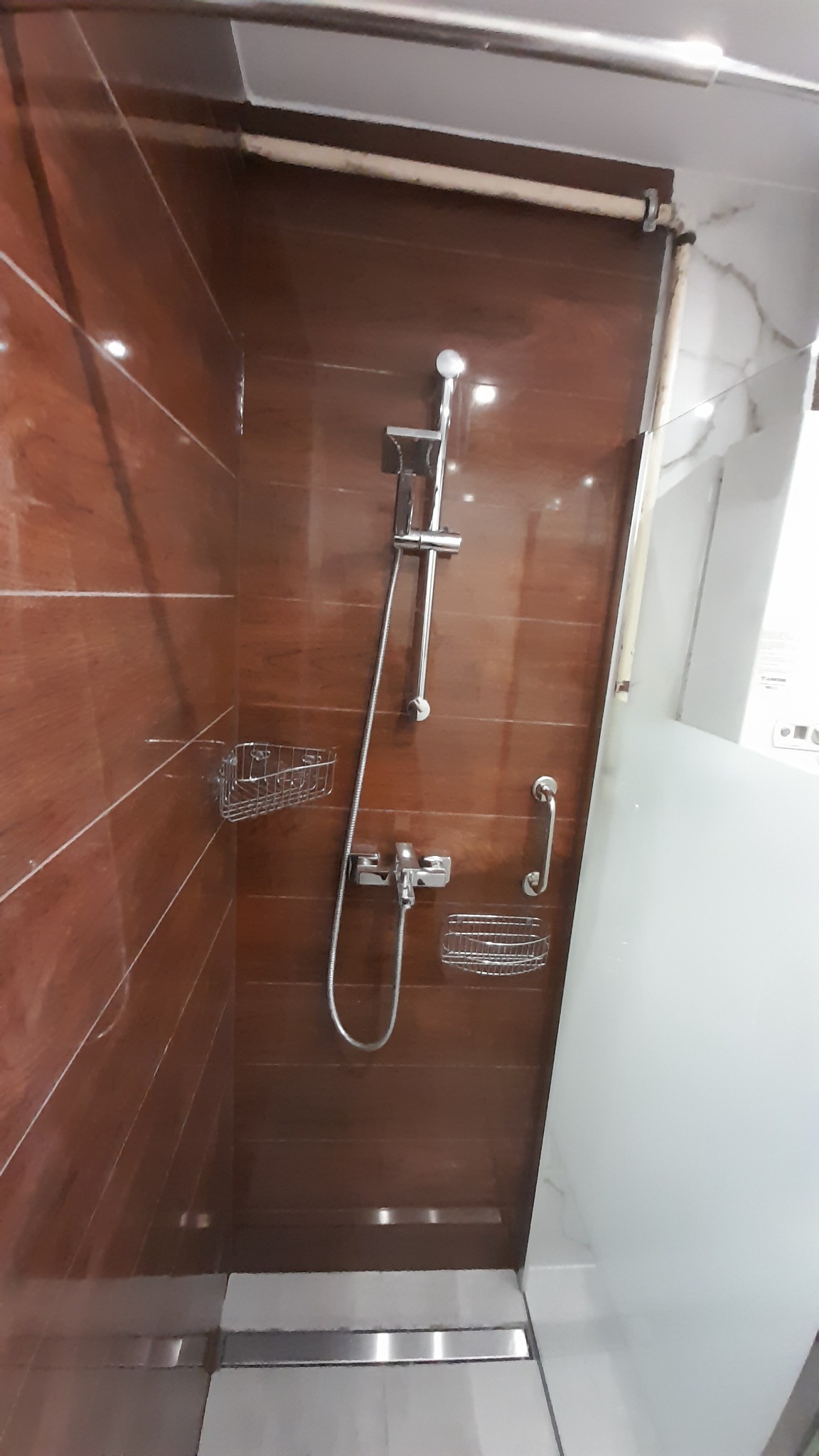 Kabina prysznicowa z brązowymi płytkami imitującymi drewno, wyposażona w deszczownicę, półki na kosmetyki i uchwyt dla niepełnosprawnych.