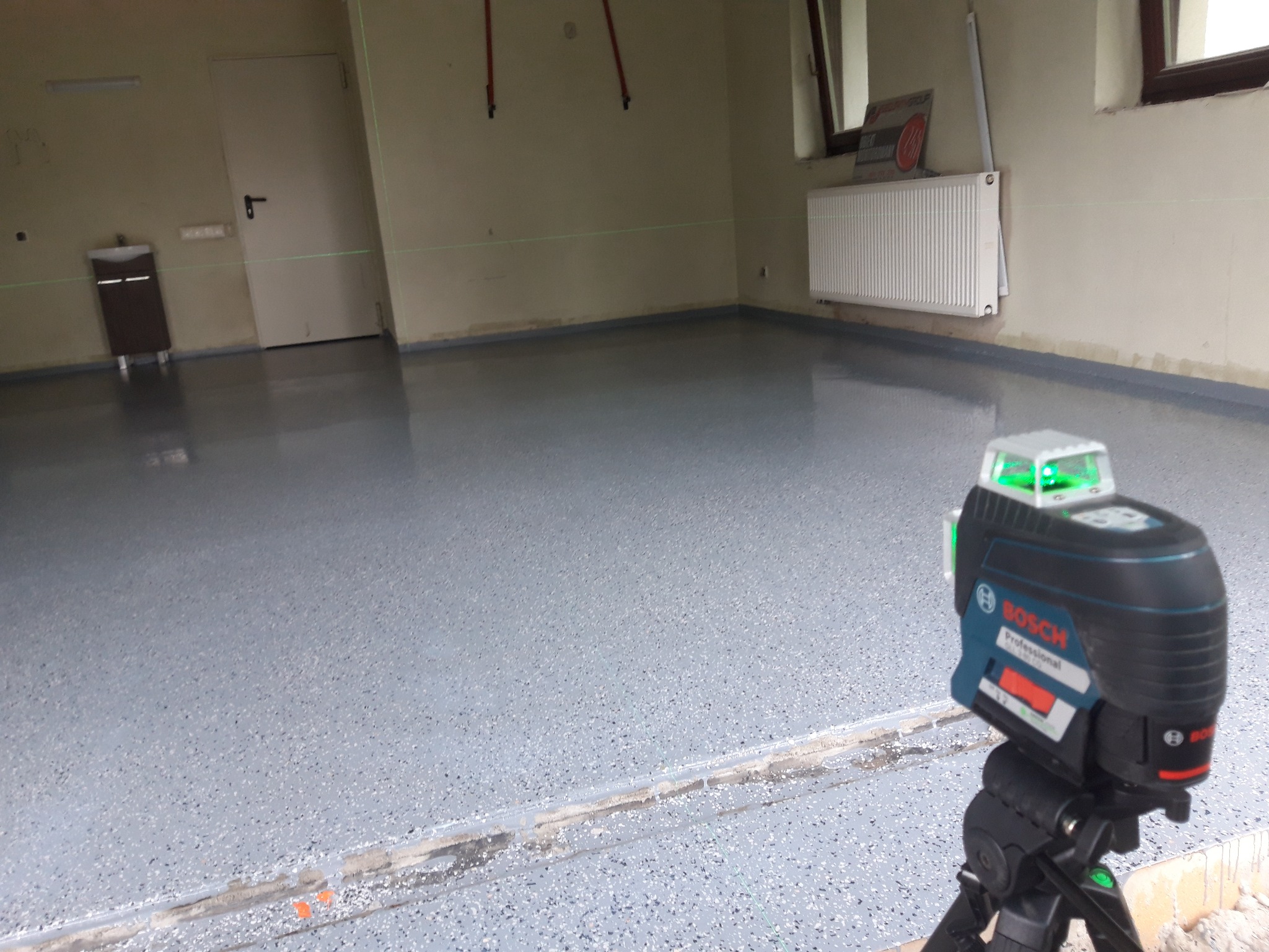 Świeżo wylana, jasnoszara posadzka w pomieszczeniu z widocznym laserowym niwelatorem Bosch na trójnogu, wyznaczającym poziom. Na ścianach widoczne ślady po pracach wykończeniowych, grzejnik i drzwi.