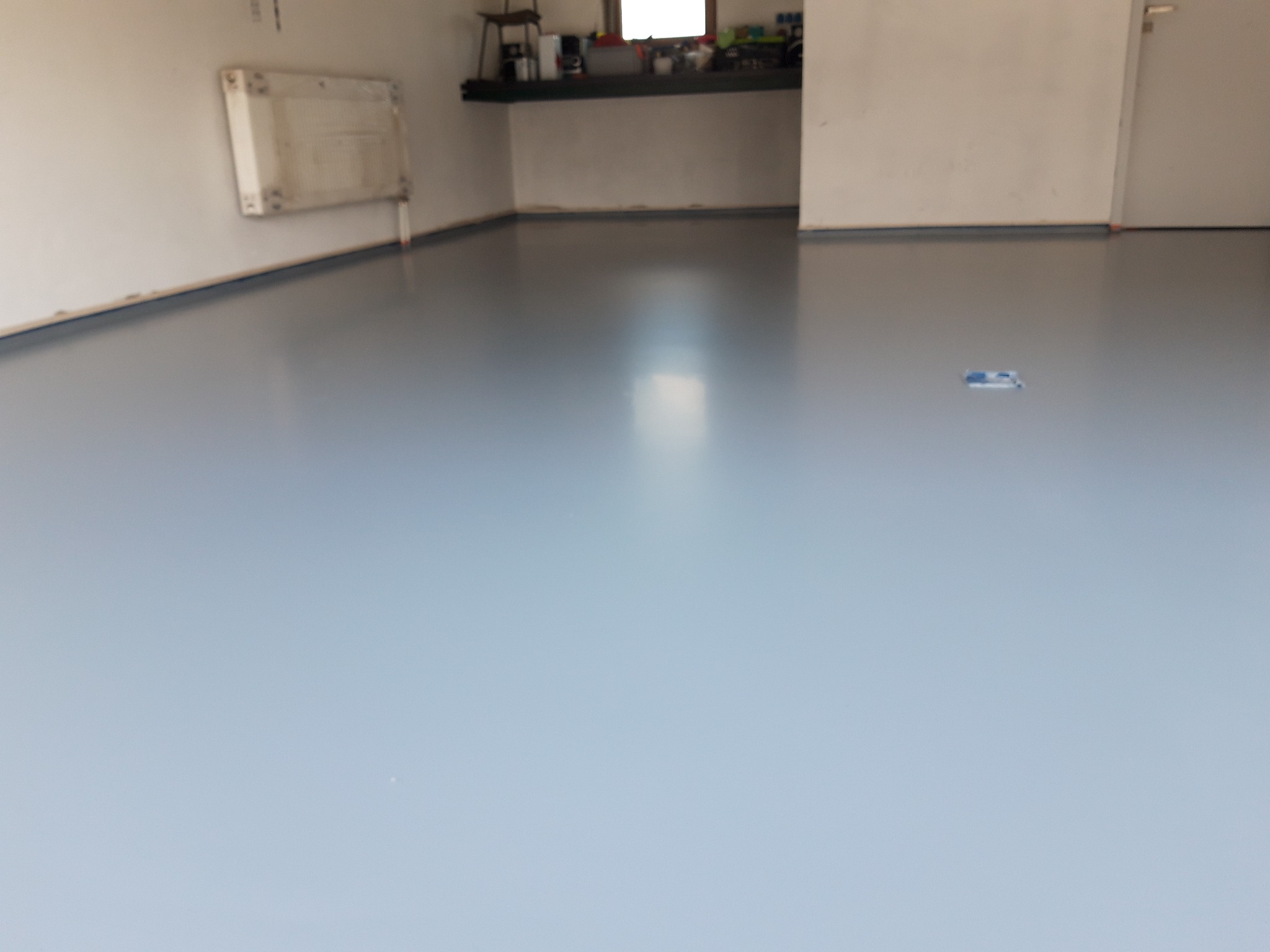 Błękitna posadzka żywiczna w garażu, widoczne ślady po taśmie malarskiej przy ścianach, półka z akcesoriami w tle.