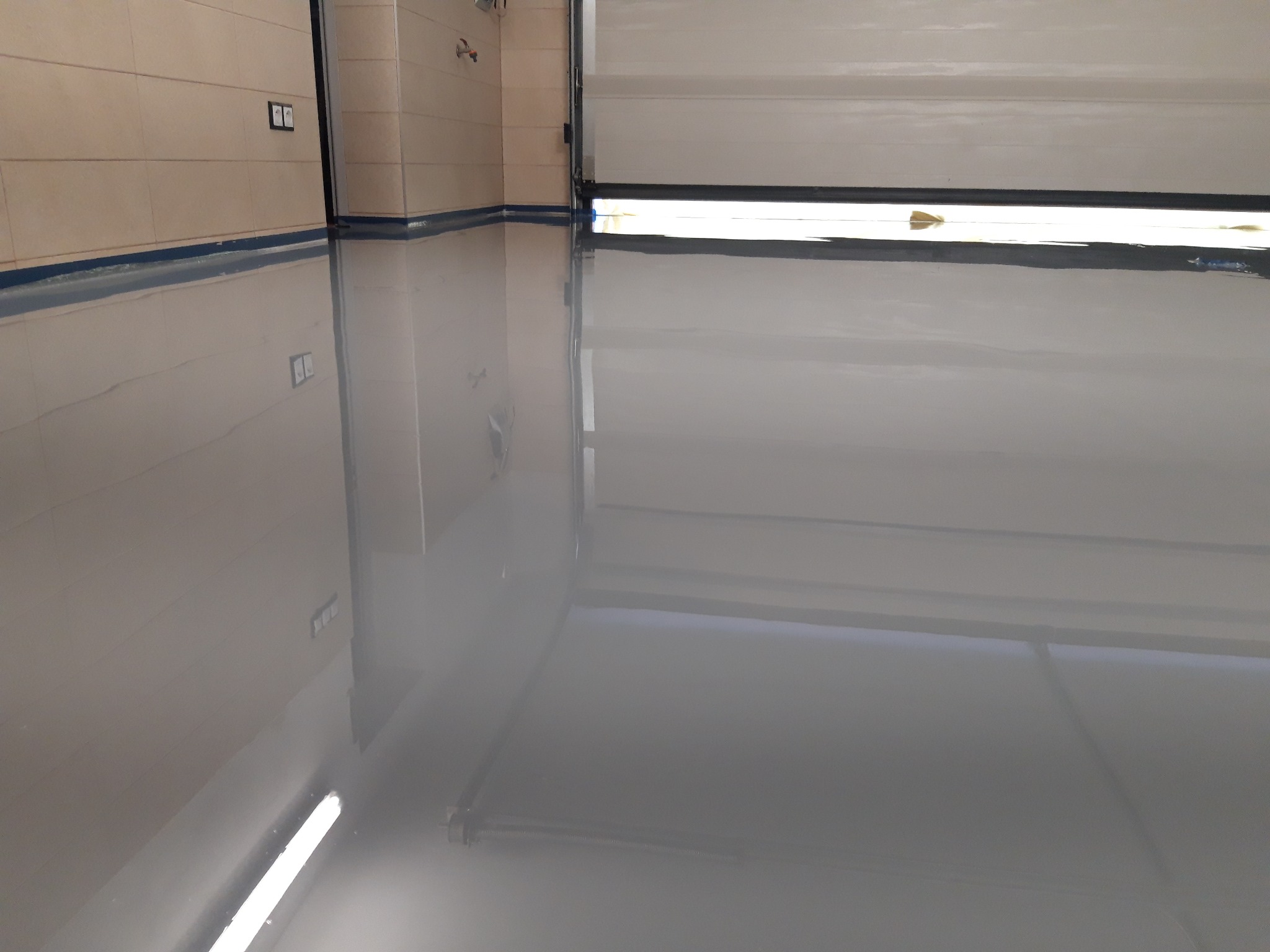 Błyszcząca, szara podłoga w garażu z widoczną bramą segmentową i panelami ściennymi, z niebieską listwą przypodłogową.