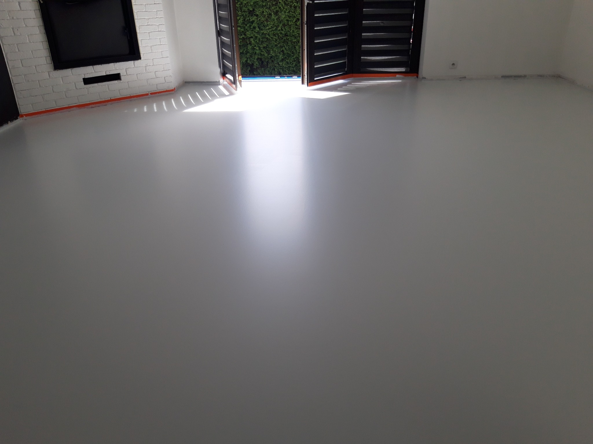 Jasnoszara, gładka posadzka żywiczna w nowym wnętrzu z ceglaną ścianą i otwartymi drzwiami na zewnątrz, wpadające światło dzienne.