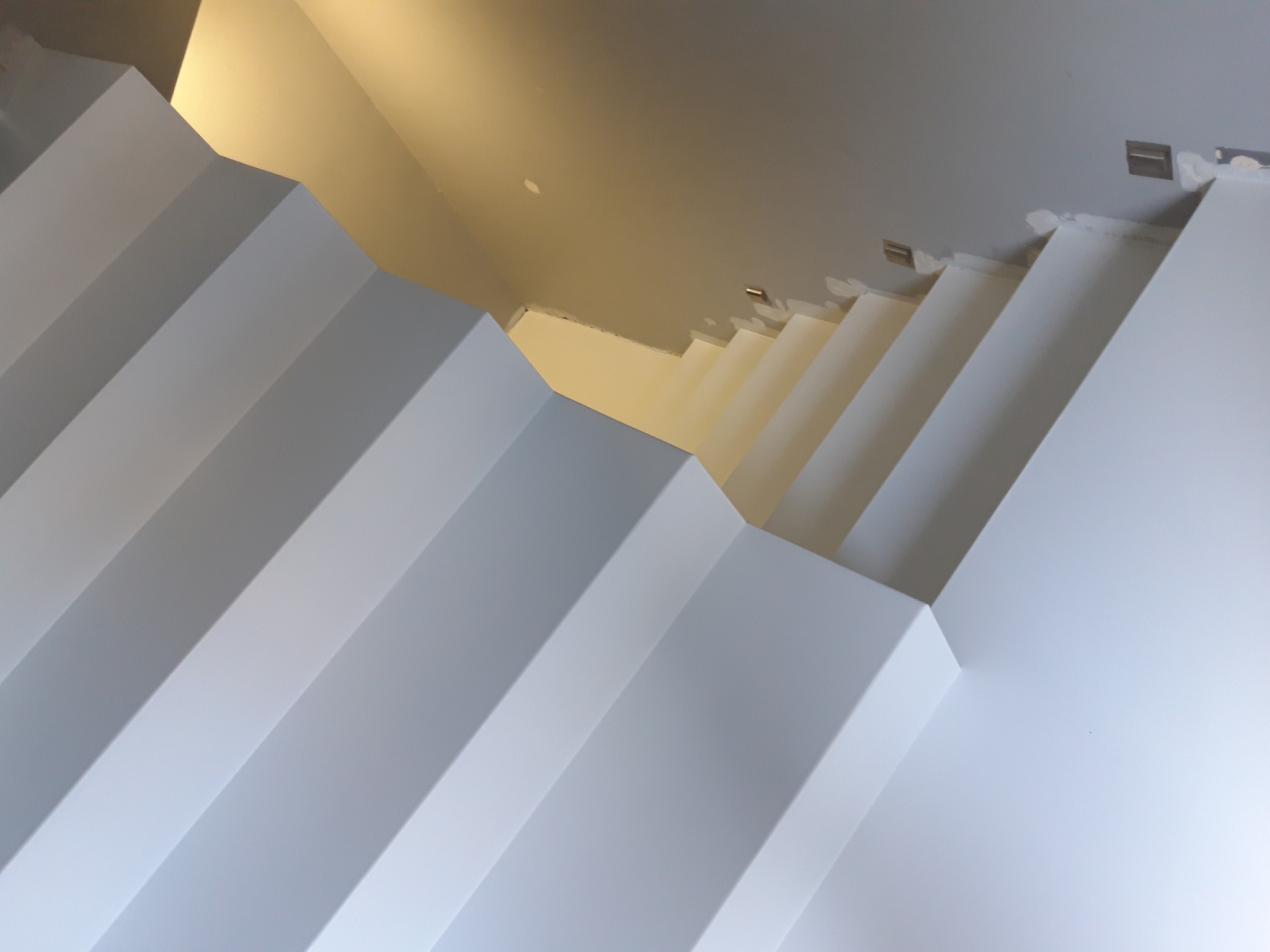 Minimalistyczne, białe schody z gładkimi powierzchniami, widziane z dołu pod kątem, z fragmentem sufitu i oświetleniem w tle. Widoczne detale wykończenia.