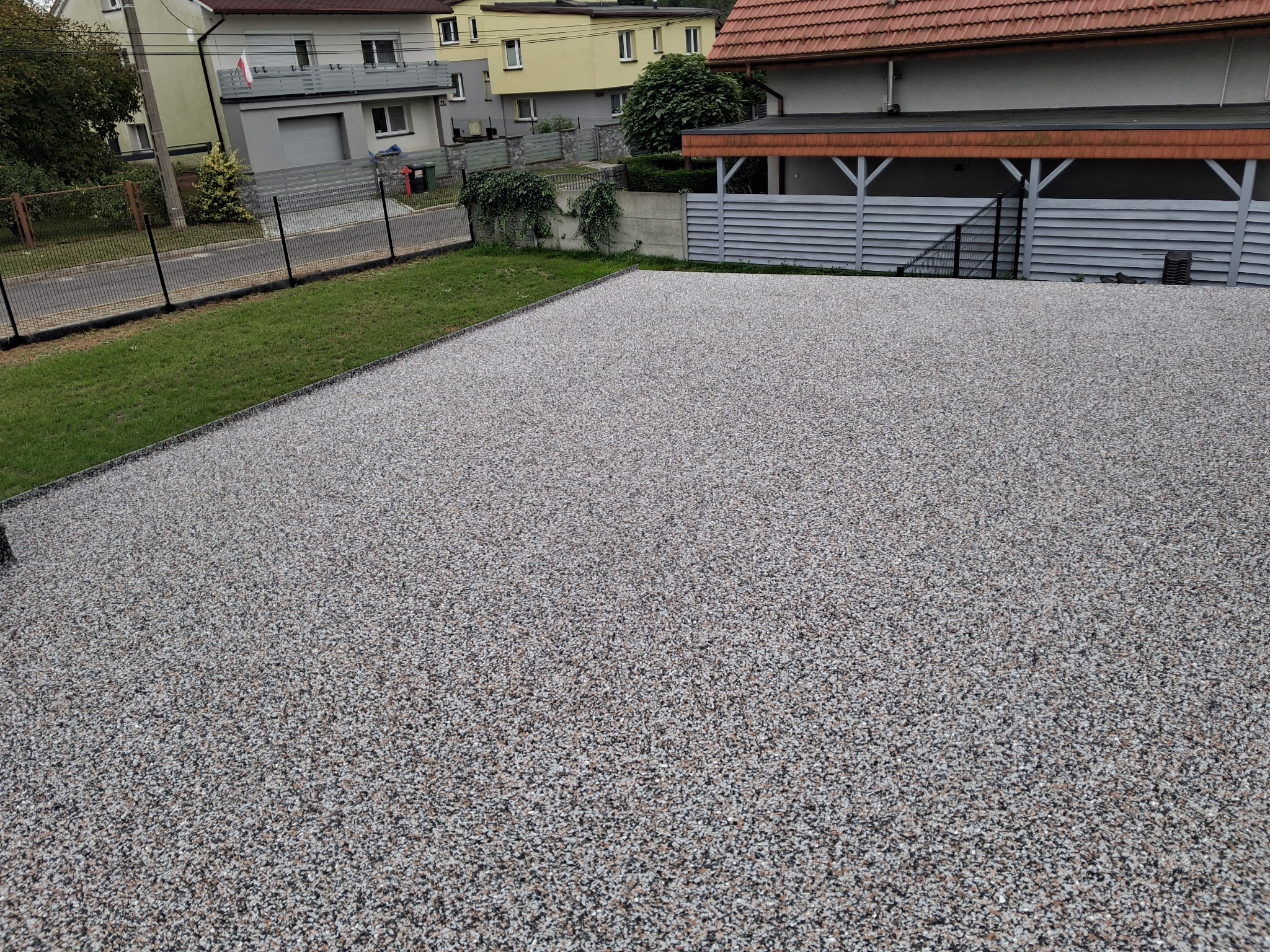 Szara, drobnoziarnista nawierzchnia z żywicy epoksydowej na podjeździe, obok trawnik i ogrodzenie. W tle zabudowa jednorodzinna, widok z góry.