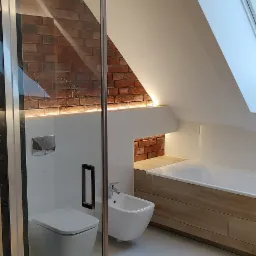 Nowoczesna łazienka na poddaszu z kabiną prysznicową, toaletą podwieszaną, bidetem i wanną obudowaną drewnem, akcentowana cegłą i oświetleniem LED.