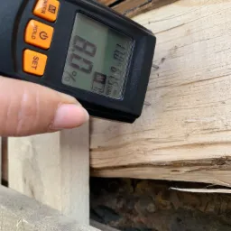 Pomiar wilgotności drewna kominkowego za pomocą wilgotnościomierza elektronicznego, wyświetlającego wynik 8.0%.