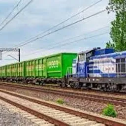 Długi pociąg towarowy z wagonami w kolorze zielonym i lokomotywą w kolorze niebieskim, widoczny na torach kolejowych na tle błękitnego nieba z chmurami i zielenią po bokach.