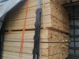 Stos jasnych, równo ułożonych desek w przestrzeni ładunkowej, zabezpieczony pomarańczowym pasem transportowym i czarną belką stabilizującą.