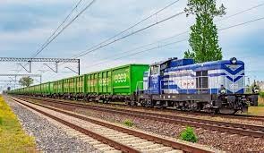 Długi pociąg towarowy z wagonami w kolorze zielonym i lokomotywą w kolorze niebieskim, widoczny na torach kolejowych na tle błękitnego nieba z chmurami i zielenią po bokach.