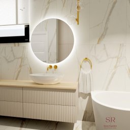 Projektowanie wnętrz Sylwia Rygielska - Elegancka łazienka z białymi marmurowymi płytkami, okrągłym lustrem podświetlanym LED, drewnianą szafką pod umywalką z frezowanymi frontami, złotą armaturą i wolnostojącą wanną.