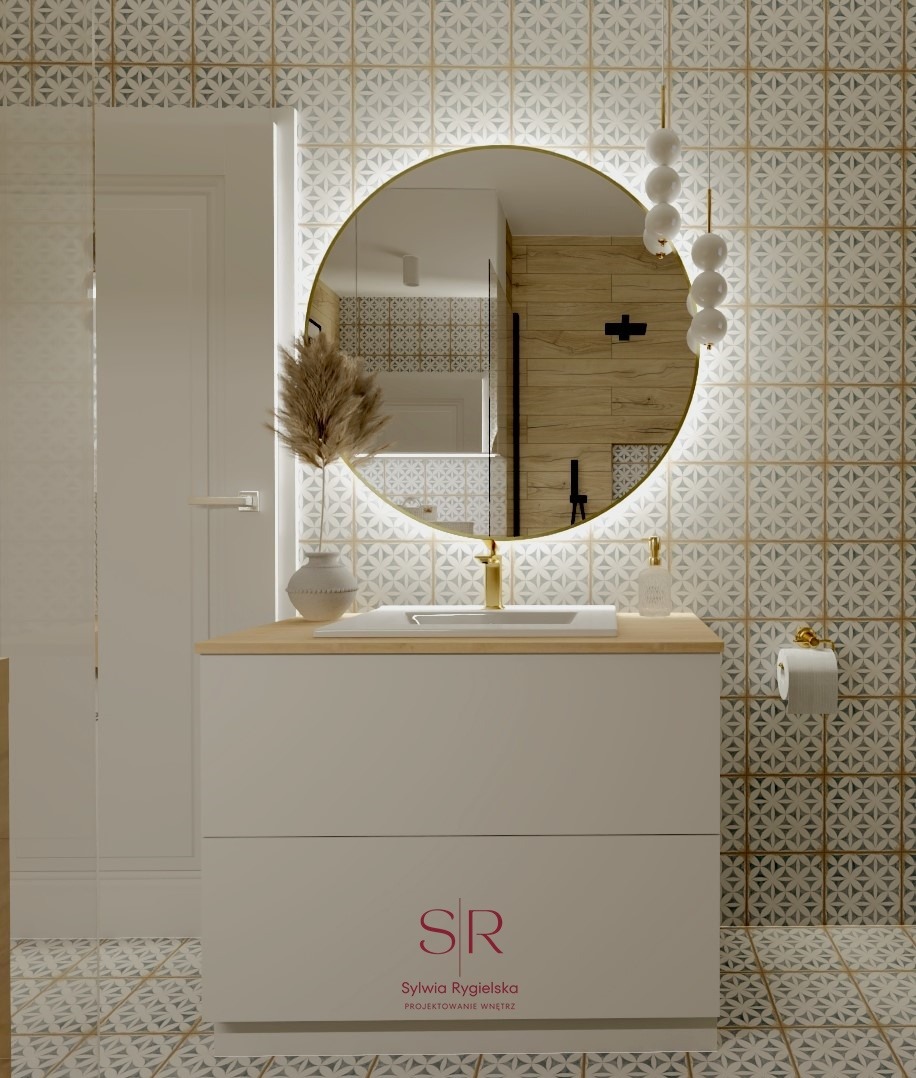 Elegancka łazienka z okrągłym lustrem podświetlanym LED, złotą armaturą i wzorzystymi płytkami na ścianach i podłodze. Na blacie umywalki dekoracyjny wazon z suszonymi trawami i dozownik do mydła.