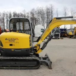 Żółta mini koparka na gąsienicach z łyżką, zaparkowana na placu z innymi maszynami budowlanymi i ciężarówkami w tle, widoczne logo wypożyczalni na korpusie.