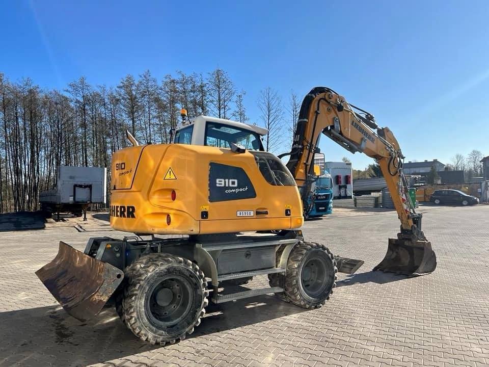 Żółta koparka kołowa Liebherr 910 Compact na placu z kostki brukowej, widoczny lemiesz z przodu, błękitne niebo i drzewa w tle.