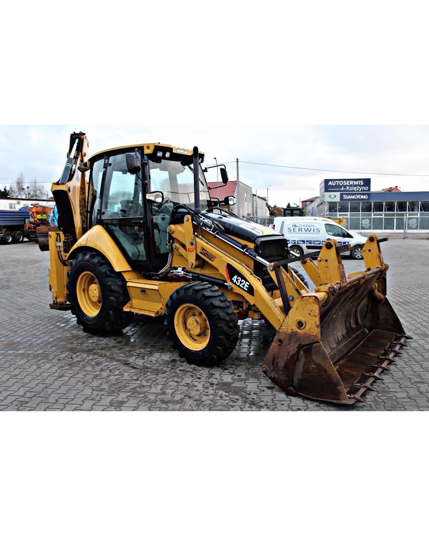 Żółta koparko-ładowarka Caterpillar 432E na brukowanym placu, z widocznymi śladami użytkowania i błota na łyżce, w tle budynek serwisu samochodowego i pochmurne niebo.