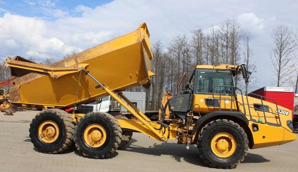 Żółty wozidło przegubowe Volvo A30E z podniesioną skrzynią ładunkową, widoczne opony z bieżnikiem, siłowniki hydrauliczne i kabina operatora na tle drzew bez liści i częściowo zachmurzonego nieba.