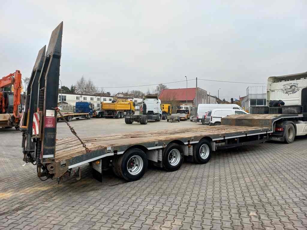 Naczepa niskopodwoziowa z rampami najazdowymi, zaparkowana na placu z kostki, w tle inne pojazdy budowlane i zabudowania.