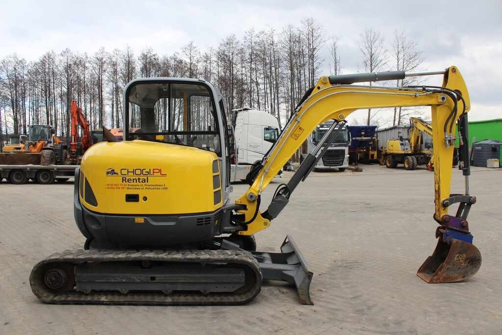 Żółta mini koparka na gąsienicach z łyżką, zaparkowana na placu z innymi maszynami budowlanymi i ciężarówkami w tle, widoczne logo wypożyczalni na korpusie.