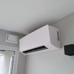 Biały klimatyzator Daikin zamontowany na szarej ścianie w pomieszczeniu, obok czarna lampka ścienna i kratka wentylacyjna, widok z dołu.