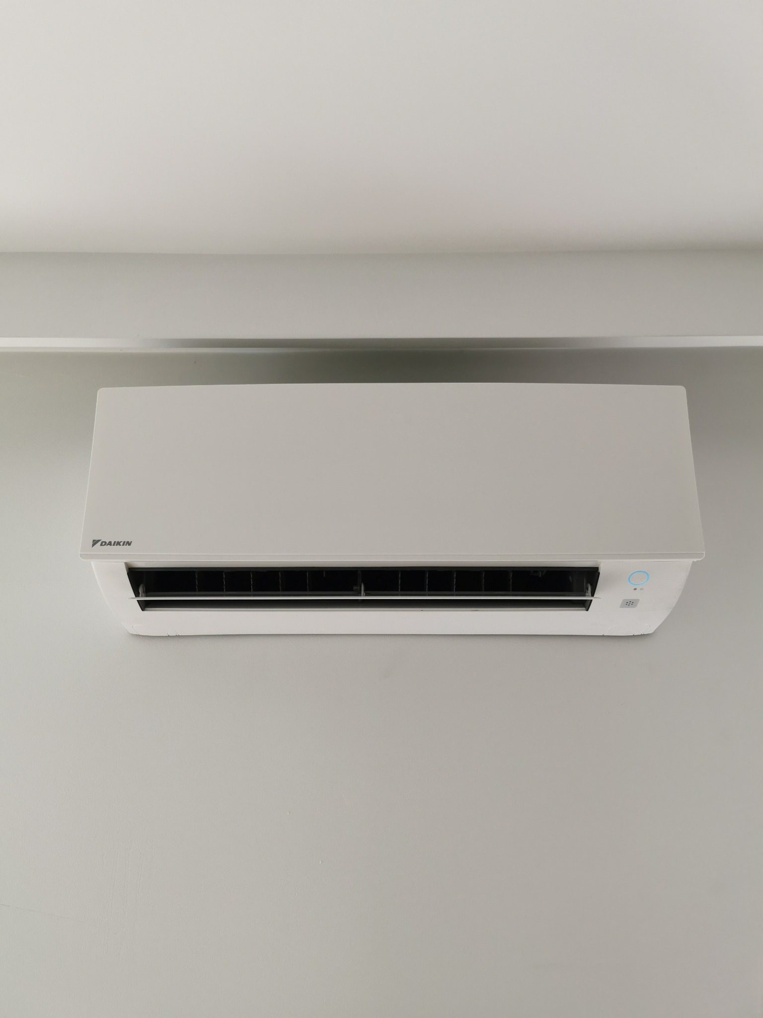 Biały klimatyzator Daikin zamontowany na szarej ścianie w pomieszczeniu z białym sufitem i listwą.