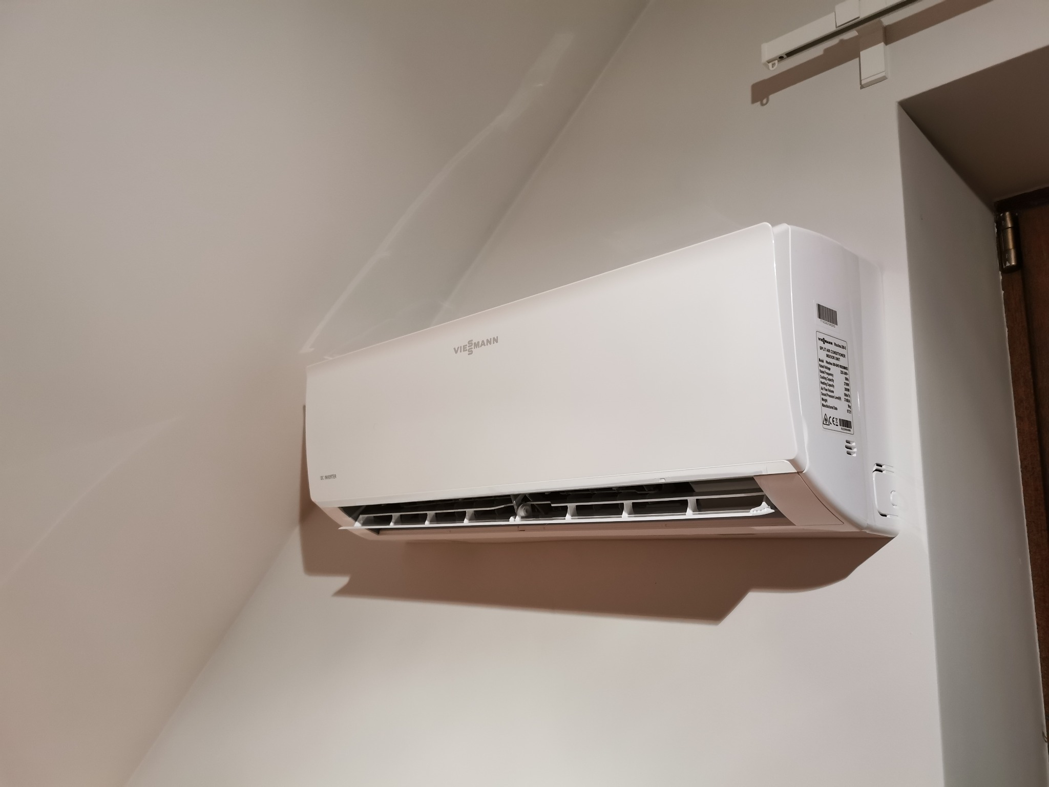 Biały klimatyzator Viessmann zamontowany na ścianie w pokoju o skośnym suficie, widoczna tabliczka znamionowa urządzenia.