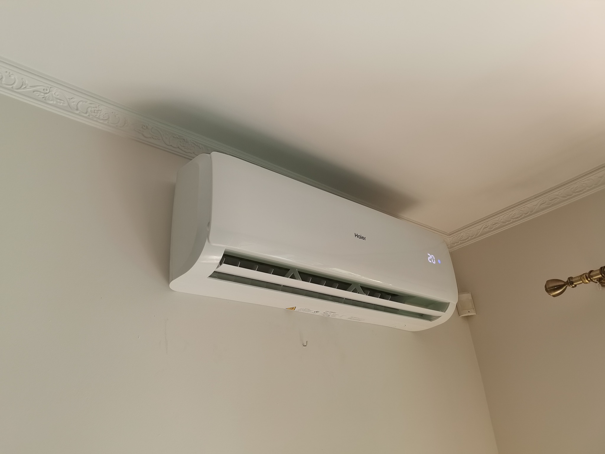 Biała klimatyzacja Haier zamontowana na ścianie w jasnym pokoju z ozdobną listwą sufitową, ustawiona na 20 stopni Celsjusza.