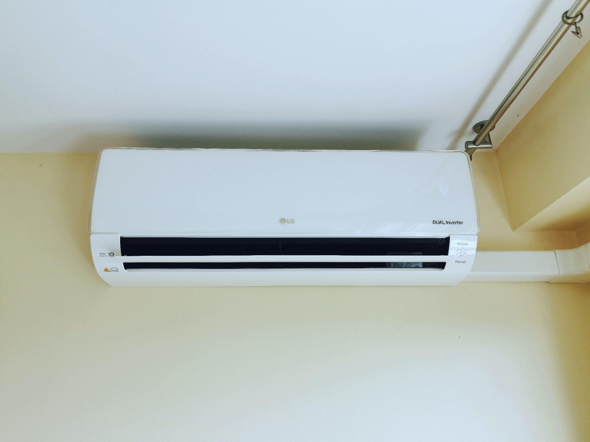 Biała jednostka wewnętrzna klimatyzatora LG Dual Inverter zamontowana na kremowej ścianie, widoczne przewody i rurki instalacyjne.