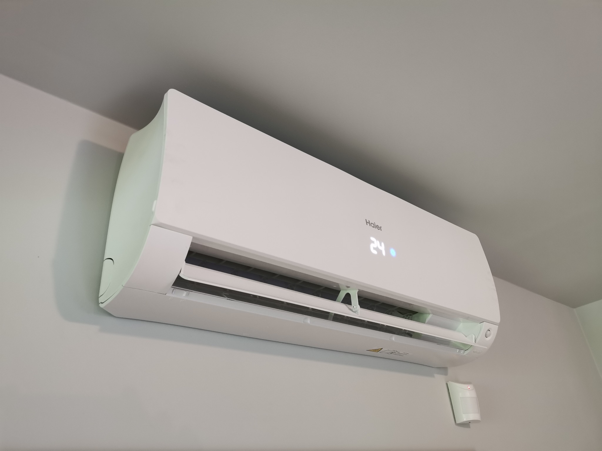Biały klimatyzator Haier zamontowany na jasnoszarej ścianie, z widocznym czujnikiem ruchu poniżej i wyświetlaczem temperatury 24 stopnie Celsjusza.